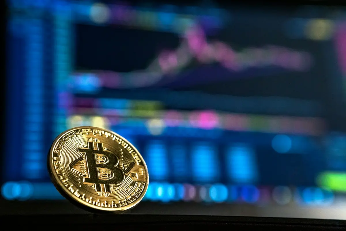 Gold bitcoin coin standing in front of a blurred trading screen showing price charts